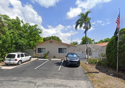 Exterior front view of Paradise Senior Residence assisted living facility in Florida, a beige stucco building with a red tile roof, palm tree, American flag, lush greenery, and private parking lot on a sunny day